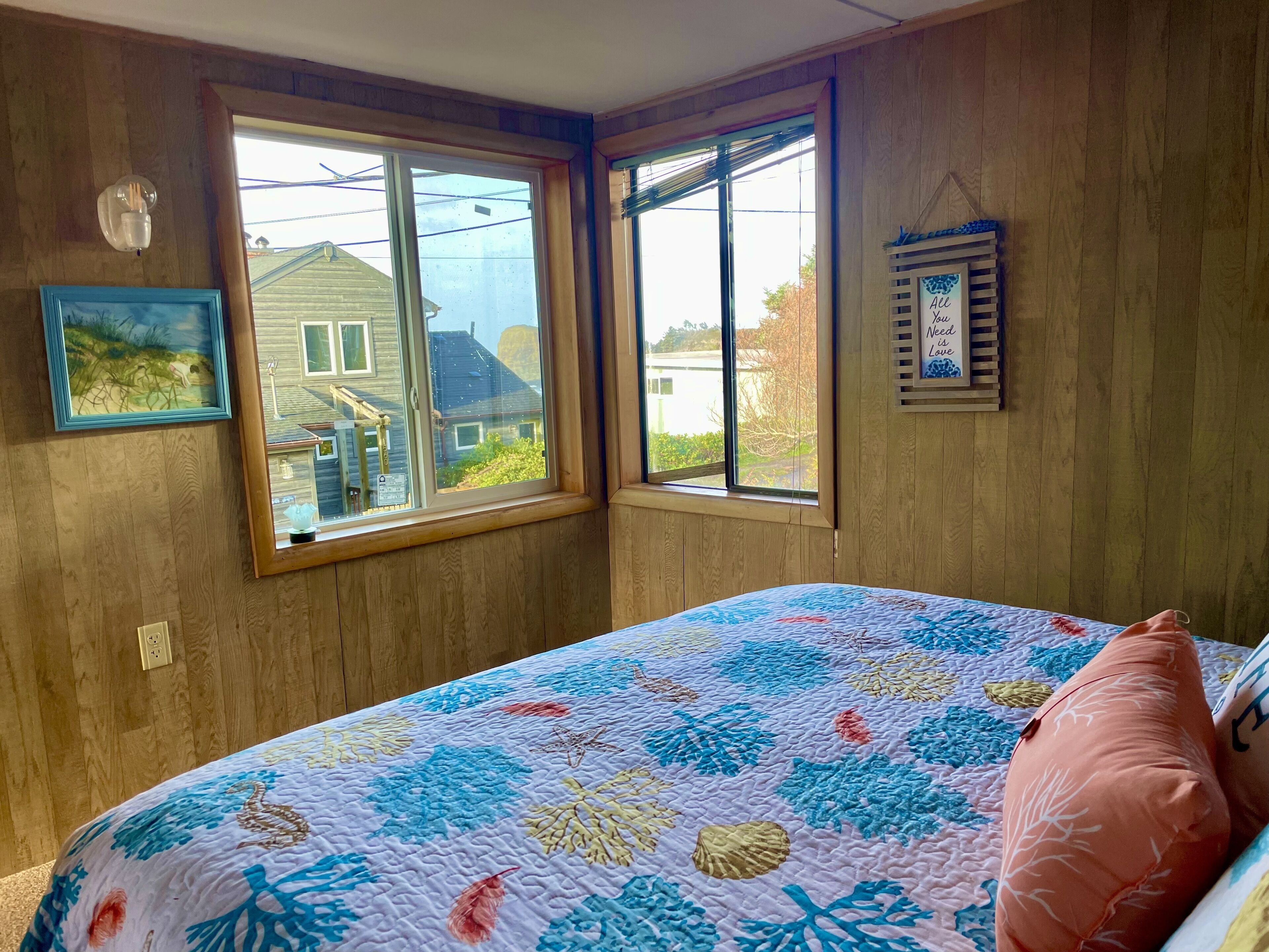 Un dormitorio con un ventanal grande, una cama con colcha floral y un estante de pared con un cuadro enmarcado.