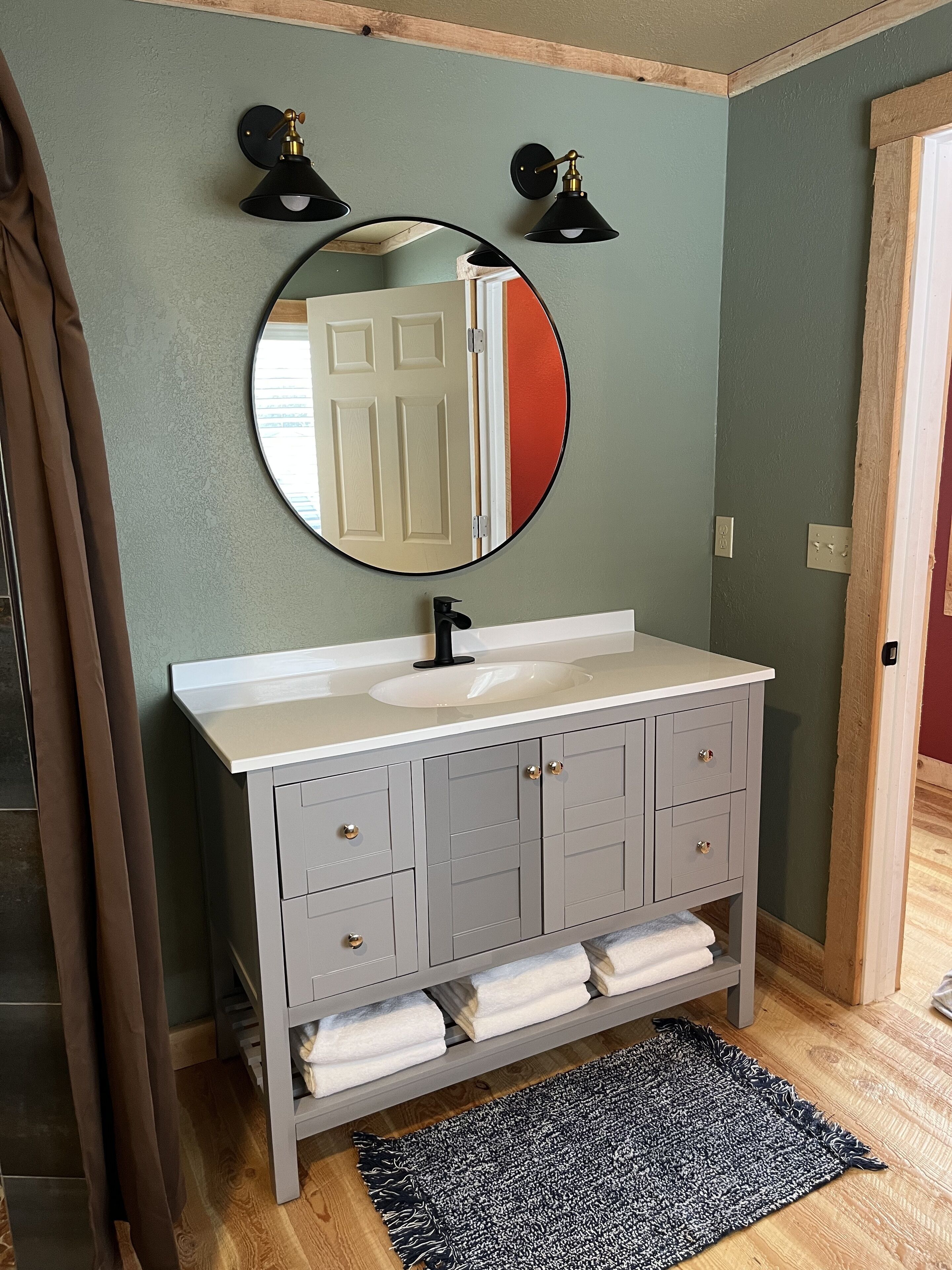 A bathroom with a round mirror, a white sink, grey cabinets, and a black and white rug.