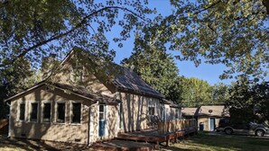 Een rustiek houten huis met een veranda en een klein terras, omringd door bomen en een heldere lucht.