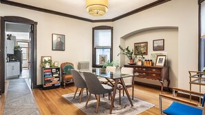 Un comedor con una mesa y sillas de vidrio, un aparador de madera con objetos decorativos, y vista a la cocina a través de una puerta abierta.
