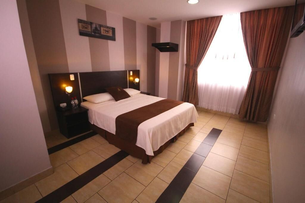 A hotel room with a large bed, two bedside tables with lamps, a headboard, a window with curtains, and a wall-mounted picture.