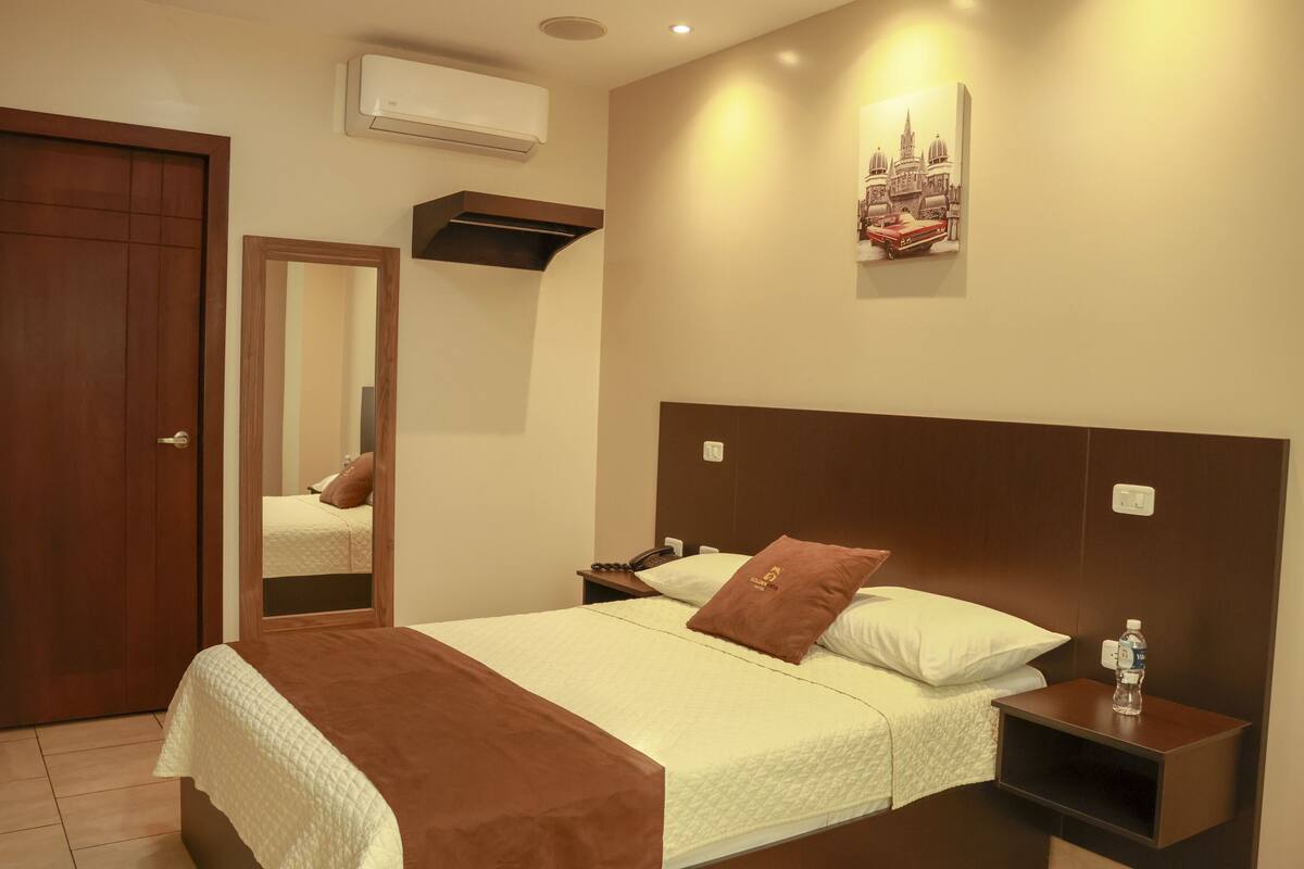 A hotel room with a bed, a nightstand, a mirror, an air conditioner, and a wall-mounted artwork.