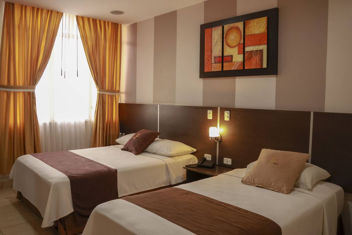 A hotel room with two beds, a large window with curtains, a wall-mounted artwork, and a bedside table with a lamp.