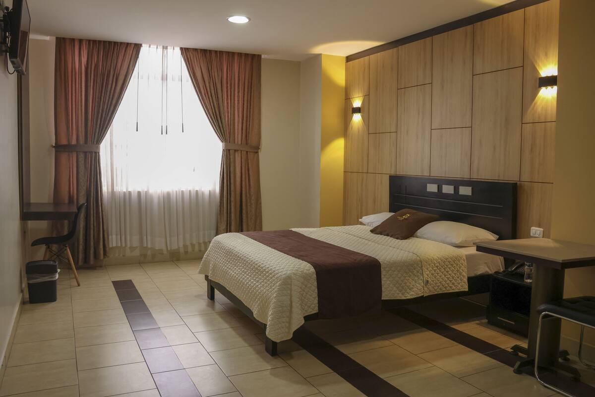 A hotel room with two beds, a desk, and a chair. There is a window with curtains, a wall-mounted light, and a wooden panel design.