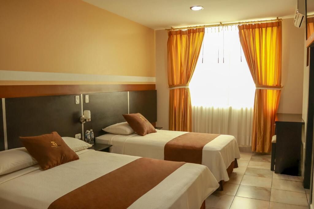 Habitación de hotel con dos camas, cabecera, mesita de noche, lámpara y ventana con cortinas.