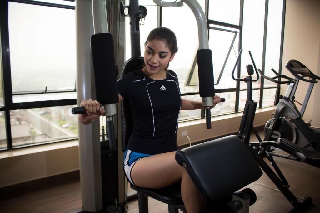 Una persona utilizando una máquina de prensa de piernas en un gimnasio con ventanas amplias y diverso equipo de ejercicio.