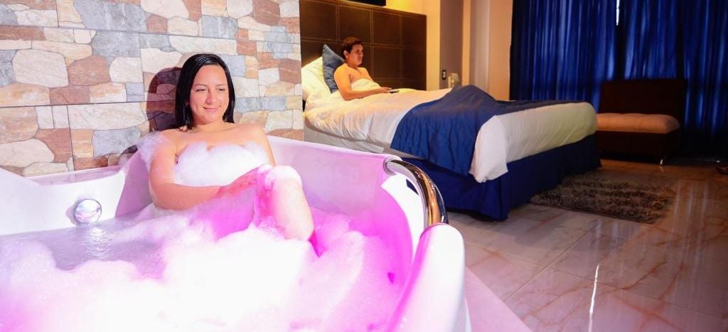 Una habitación con pared de piedra, una cama con colcha azul y una bañera llena de burbujas rosadas.