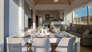A modern dining area with a white table set for four, a kitchen in the background, and a large window with a view of the landscape. - Kladissos Family Seaside Villa with heated pool, hydromassage and barbecue! (Agia Pelagia)