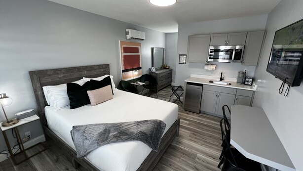 A modern hotel room with a large bed, a desk, a kitchenette, and a TV.