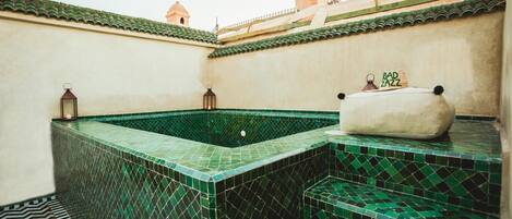 A rooftop swimming pool with green mosaic tiles, a cushioned bench, and lanterns.