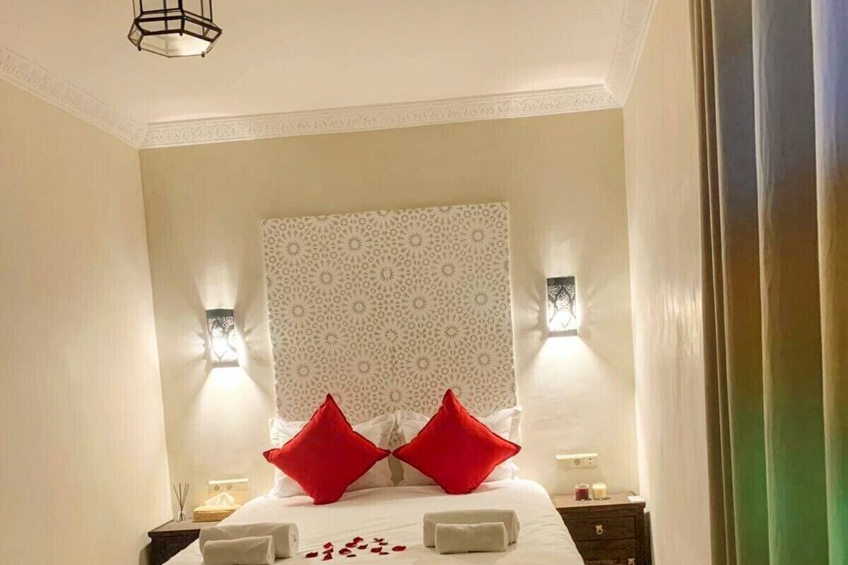 Cama bem arrumada com colcha branca e duas almofadas vermelhas, cabeceira estampada, duas luminárias de cabeceira e tapete listrado sobre piso em padrão herringbone.