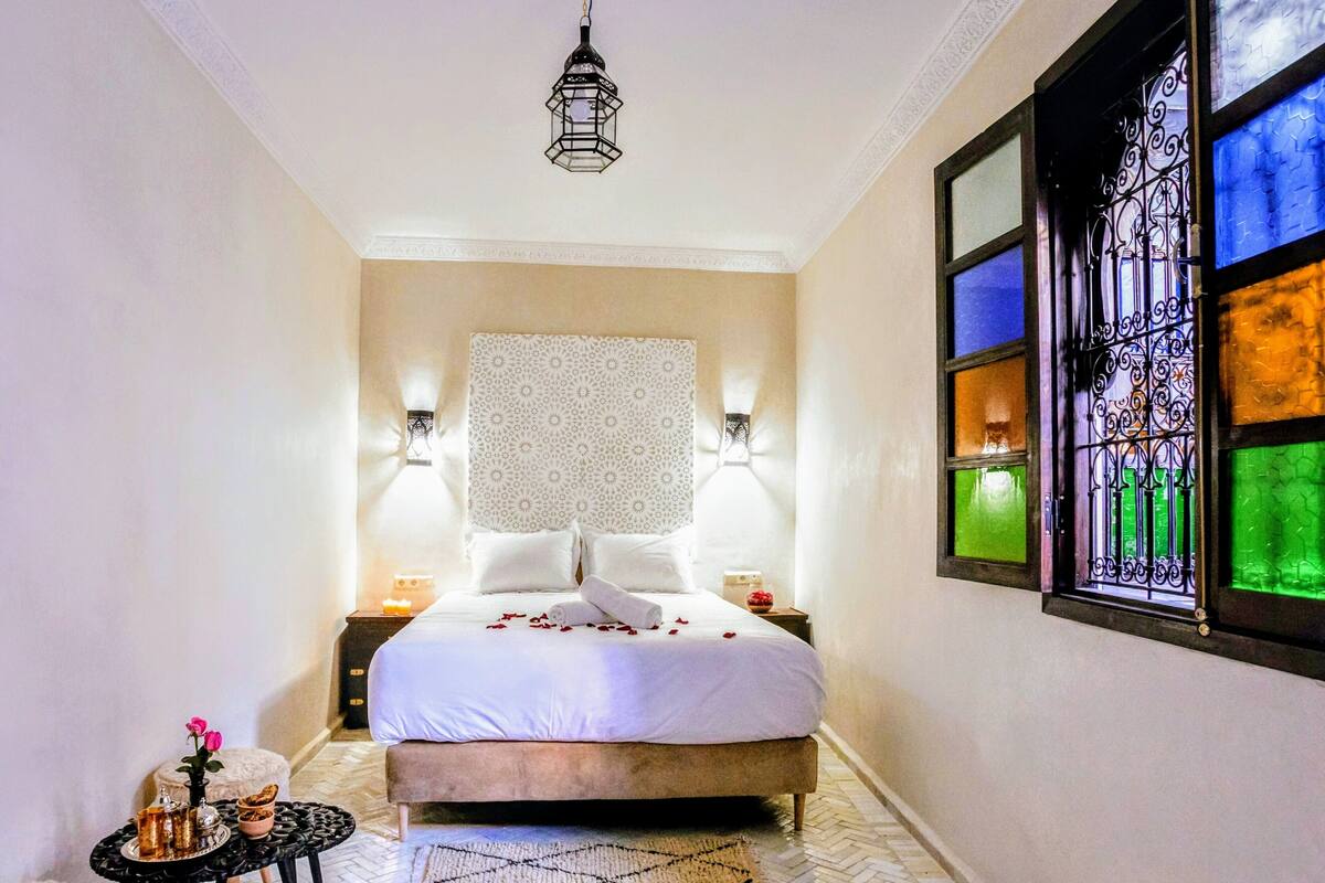 Quarto com cama, mesa de cabeceira, pequena mesa com vaso e flores, tapete decorativo e janela de vitral.
