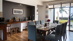 A modern dining area with a wooden table set for four, grey upholstered chairs, a coffee station, and a large window offering a view of the outdoors.