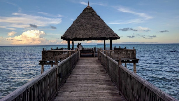 A wooden pier leading to a thatched-roof structure with a view of the sea.