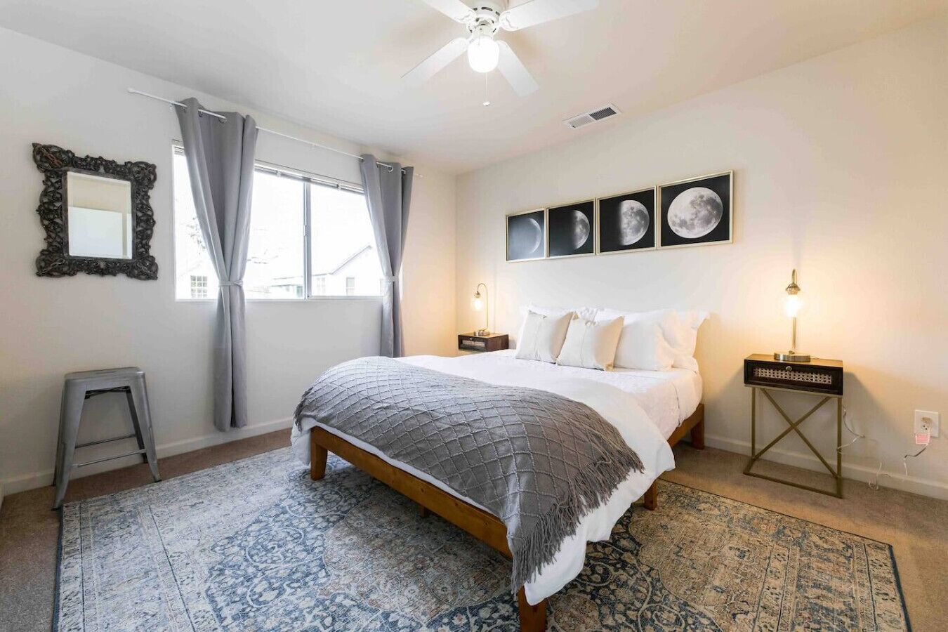 A bedroom with a bed, a nightstand, a lamp, a ceiling fan, a window with curtains, a mirror, and framed pictures on the wall.