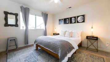 A bedroom with a bed, a nightstand, a lamp, a ceiling fan, a window with curtains, a mirror, and framed pictures on the wall.