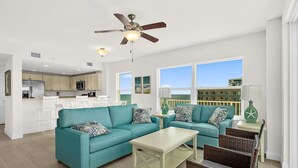 A spacious living room with teal sofas, a coffee table, and wicker chairs. A ceiling fan and a TV are visible in the background. - Beachview Luxury Condo Decks & Views (Daytona Beach)