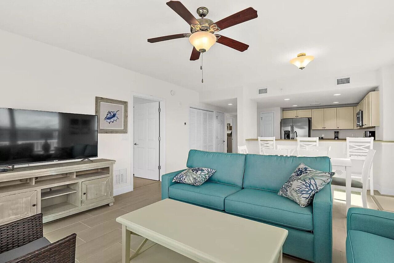 A living room with a teal sofa, a flat-screen TV, a ceiling fan, and a dining area with white chairs and a table.