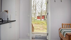 Een kamer met een bed, een houten bankje, een witte deur naar buiten en een keuken met een wit kastje en een witte bak.