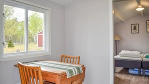 Een eetruimte met een houten tafel en stoelen, een raam met uitzicht op een rode schuur en bomen, en een slaapkamer met een bed en een lamp.