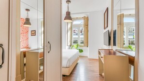 A bedroom with a bed, bedside table, dresser, and a TV. There is a window with a view of buildings outside.