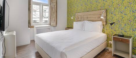 A bedroom with a bed, a bedside table, a lamp, a radiator, and a window with a view of buildings.