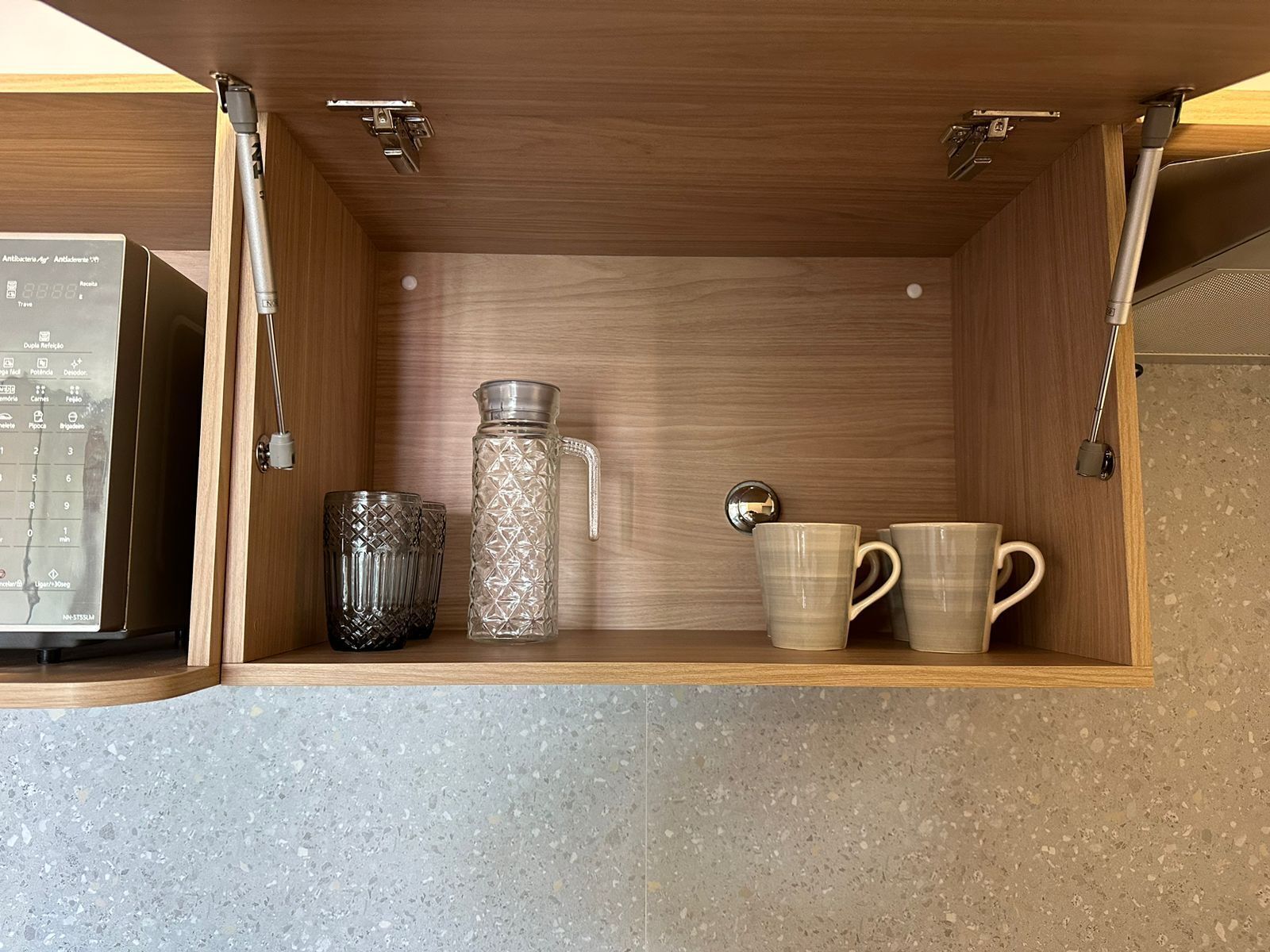 A kitchen cabinet with a microwave, a glass pitcher, and two mugs.