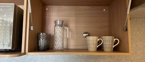 A kitchen cabinet with a microwave, a glass pitcher, and two mugs.