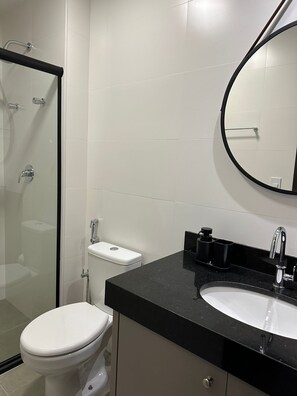 A modern bathroom with a glass shower enclosure, a white toilet, a black countertop with a sink, and a round mirror.