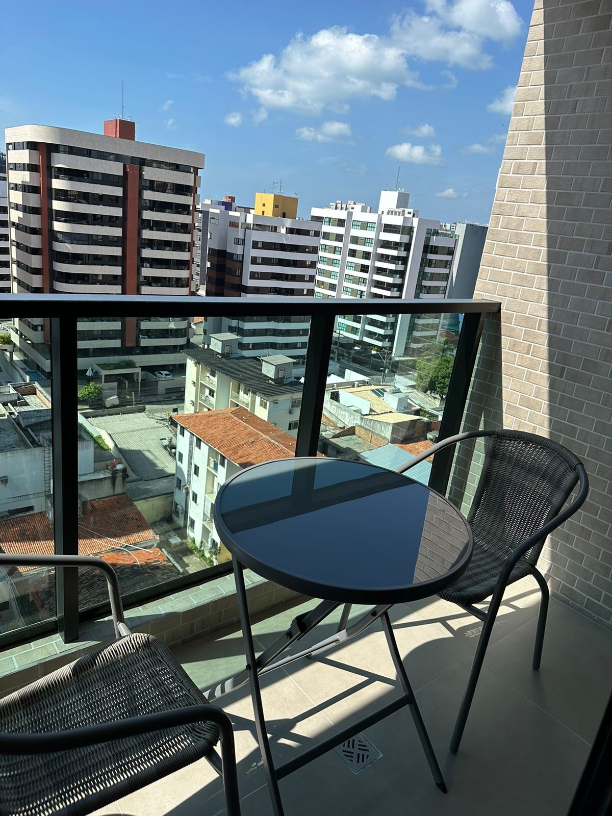 Un balcon doté d’une table en verre et de deux fauteuils en osier, offrant une vue sur un paysage urbain avec des bâtiments modernes.