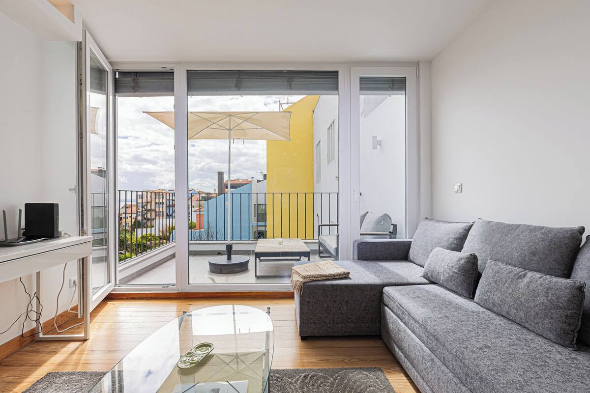A modern living room with a grey sofa, a glass coffee table, and a balcony with a yellow building in the background.