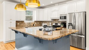 Una cucina moderna con mobili bianchi, un'isola grande con piano in granito, elettrodomestici in acciaio inossidabile e illuminazione a sospensione.