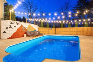 A swimming pool with lounge chairs, surrounded by a wooden fence and string lights. - Resort-style home w/ heated pool, hot tub, arcade (Fletcher)