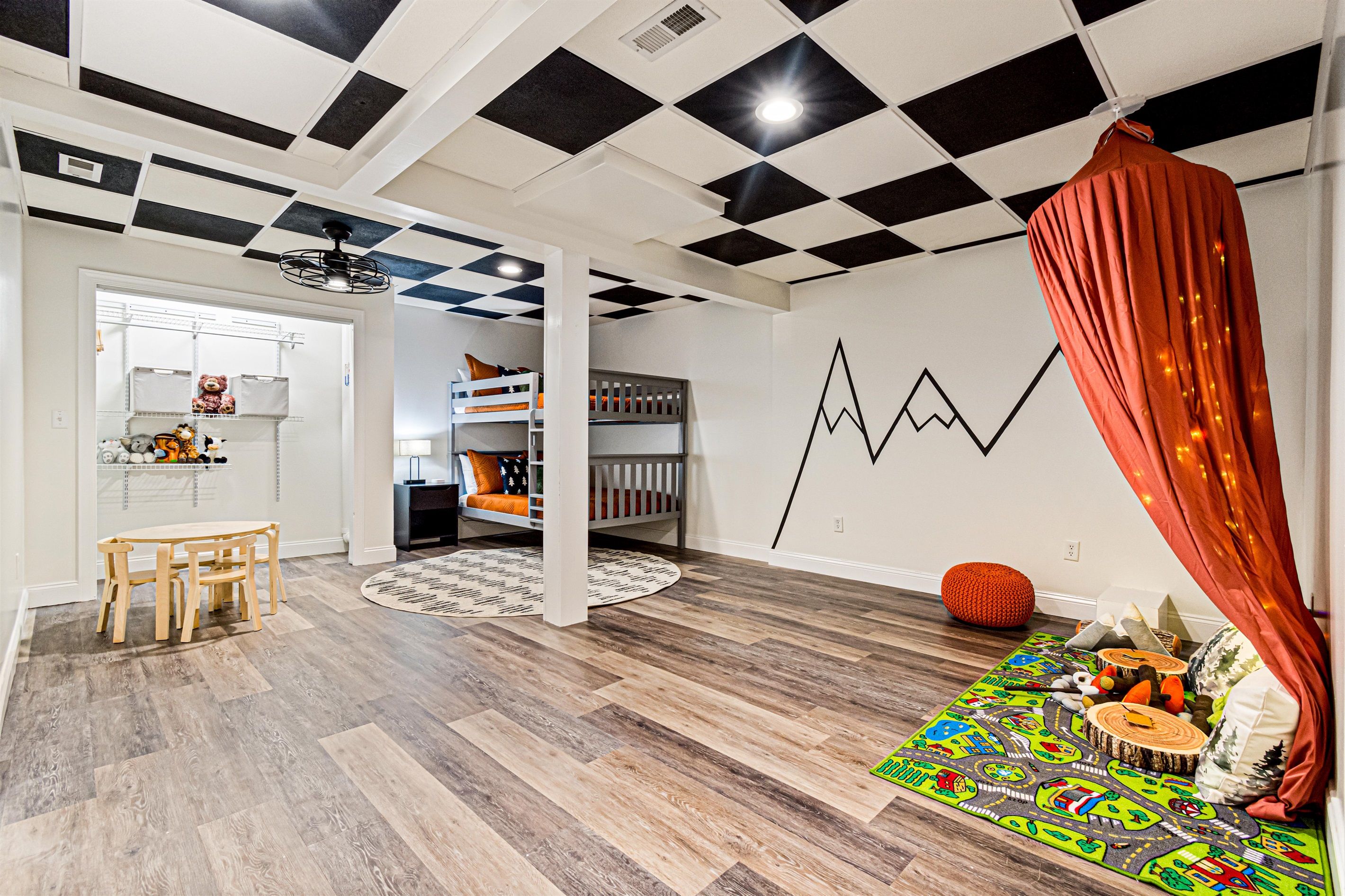 Una habitación con techo a cuadros, una litera, una zona de juegos con alfombra colorida y una mesa de comedor.