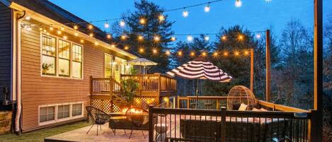 Una terraza de madera con mobiliario exterior, luces colgantes y un espacio cubierto para sentarse.