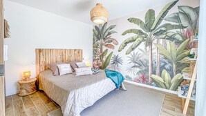 A bedroom with a large bed, a wooden headboard, a wall mural of tropical plants, a small bedside table with a lamp, and a cat climbing a shelf. - Gorgeous home in Rochegude with private swimming pool, can be inside or outside (Rochegude)
