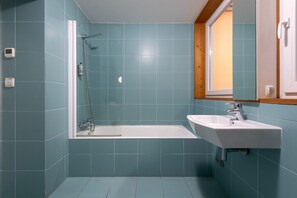 Une salle de bain avec des carreaux de couleur turquoise, une baignoire blanche et un lavabo blanc, surmonté d’un cadre de fenêtre en bois.