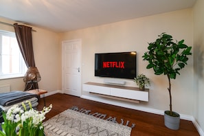 Un salon avec un téléviseur à écran plat fixé au mur, une plante en pot, une fenêtre avec des rideaux, un canapé et un tapis à motifs.