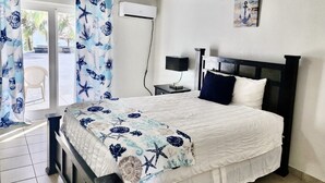 Un dormitorio con cama, ventilador de techo, equipo de aire acondicionado, mesita de noche con lámpara y vista al exterior a través de una puerta corredera.