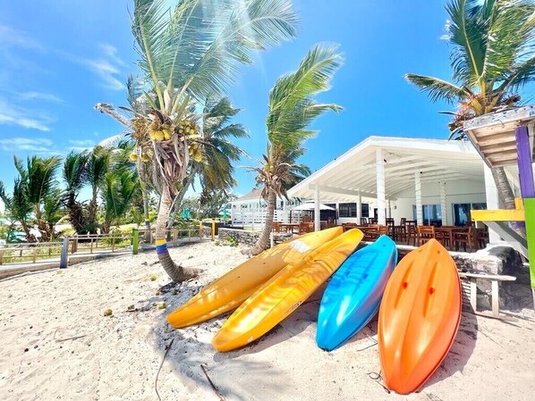 A beach with several kayaks lined up near a covered seating area with palm trees in the background.
