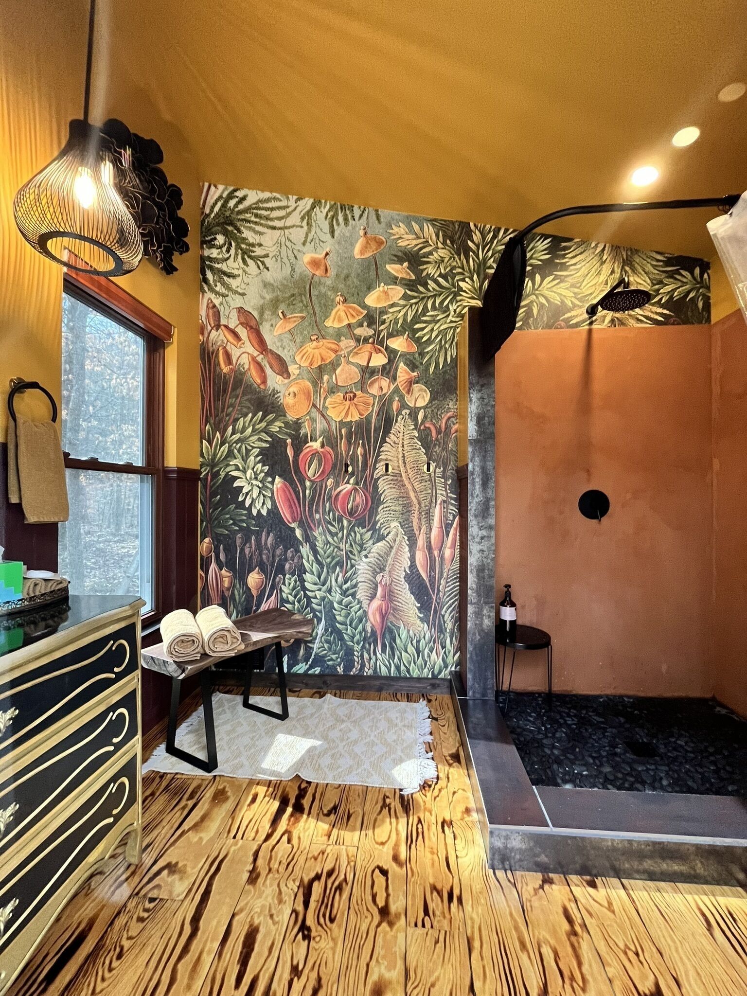 Ein Zimmer mit Holzboden, einer großen Blumenmalerei, einer Bank, einem Tisch mit Handtüchern und einer hängenden Leuchte.