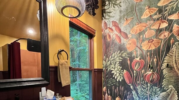Een kamer met een grote spiegel, een houten dressoir, een bankje, een wandtapijt met bloemenmotieven en een raam met uitzicht op groen.