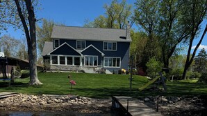 A deck with two white Adirondack chairs facing a house with a stone chimney, a grassy yard, and a dock extending into a body of water.