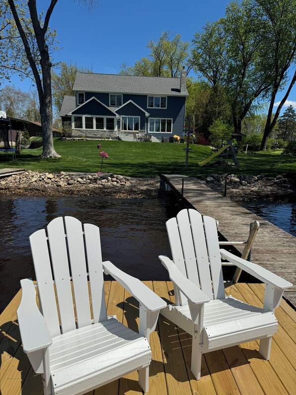 Une terrasse avec deux chaises Adirondack blanches, donnant sur une maison dotée d’une cheminée en pierre, un jardin verdoyant et une jetée s’avançant dans l’eau.
