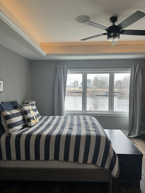 Une chambre à coucher avec un lit, un ventilateur de plafond, une fenêtre avec des rideaux et une vue sur une étendue d’eau.