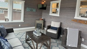 A cozy living room with a wooden ceiling, a television mounted on the wall, and a window overlooking a backyard.