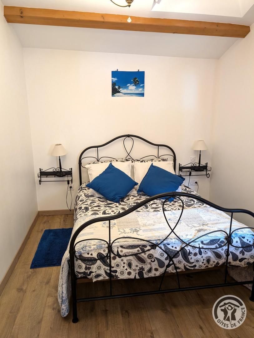 A single bed with a patterned bedspread and two blue pillows, flanked by two bedside tables with lamps, in a room with wooden beams on the ceiling and a small painting on the wall.