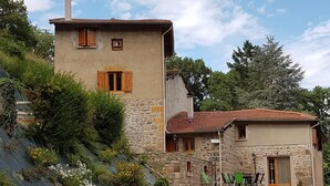 Un bâtiment en pierre avec un toit en pente, un petit porche avec des chaises et un jardin agrémenté de plantes et de fleurs.