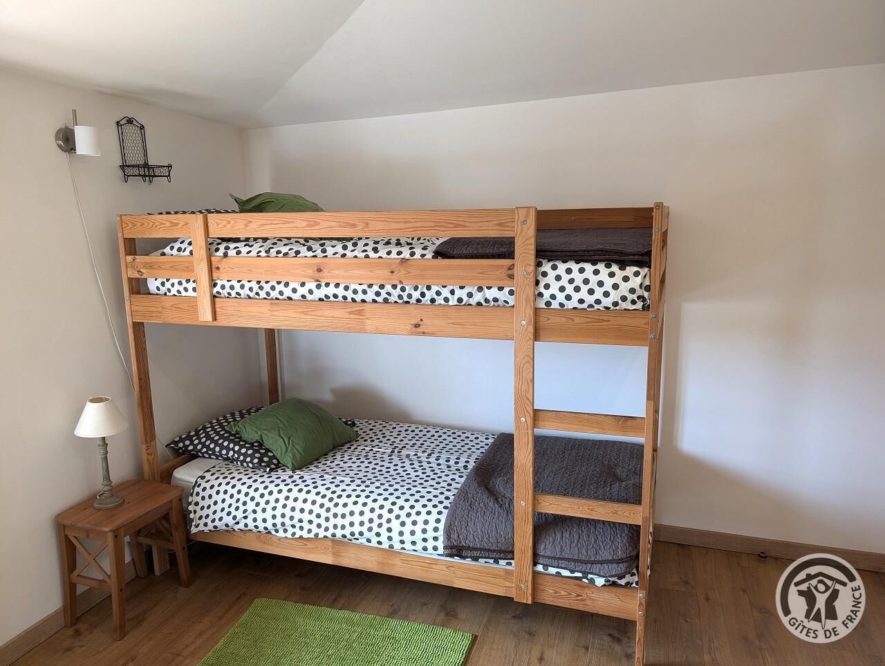 Een stapelbed met een stippenbed overtrek, een klein houten nachtkastje met een lamp, en een groen tapijt op de vloer.