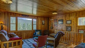 L’intérieur chaleureux d’un chalet avec des murs en bois, une cheminée, un canapé, un fauteuil et une vue sur la forêt par de grandes fenêtres.